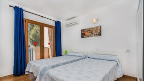 A room with two beds, a window with curtains, an air conditioning unit, and a wall-mounted artwork. - Sunset Villa Isleta (Alcúdia)
