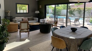 Uma sala de estar moderna com um sofá, cadeiras, uma mesa de centro e uma mesa de jantar com cadeiras. Há uma janela grande com vista para uma piscina e vegetação lá fora.