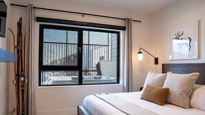 A bedroom with a large window, a bed with pillows, a bedside table, and a ski rack. - Altitude Adjustment Revelstoke Vacations (Revelstoke)