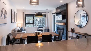 A modern living room with a fireplace, a dining area with a wooden table, and a large window offering a city view. - Altitude Adjustment Revelstoke Vacations (Revelstoke)