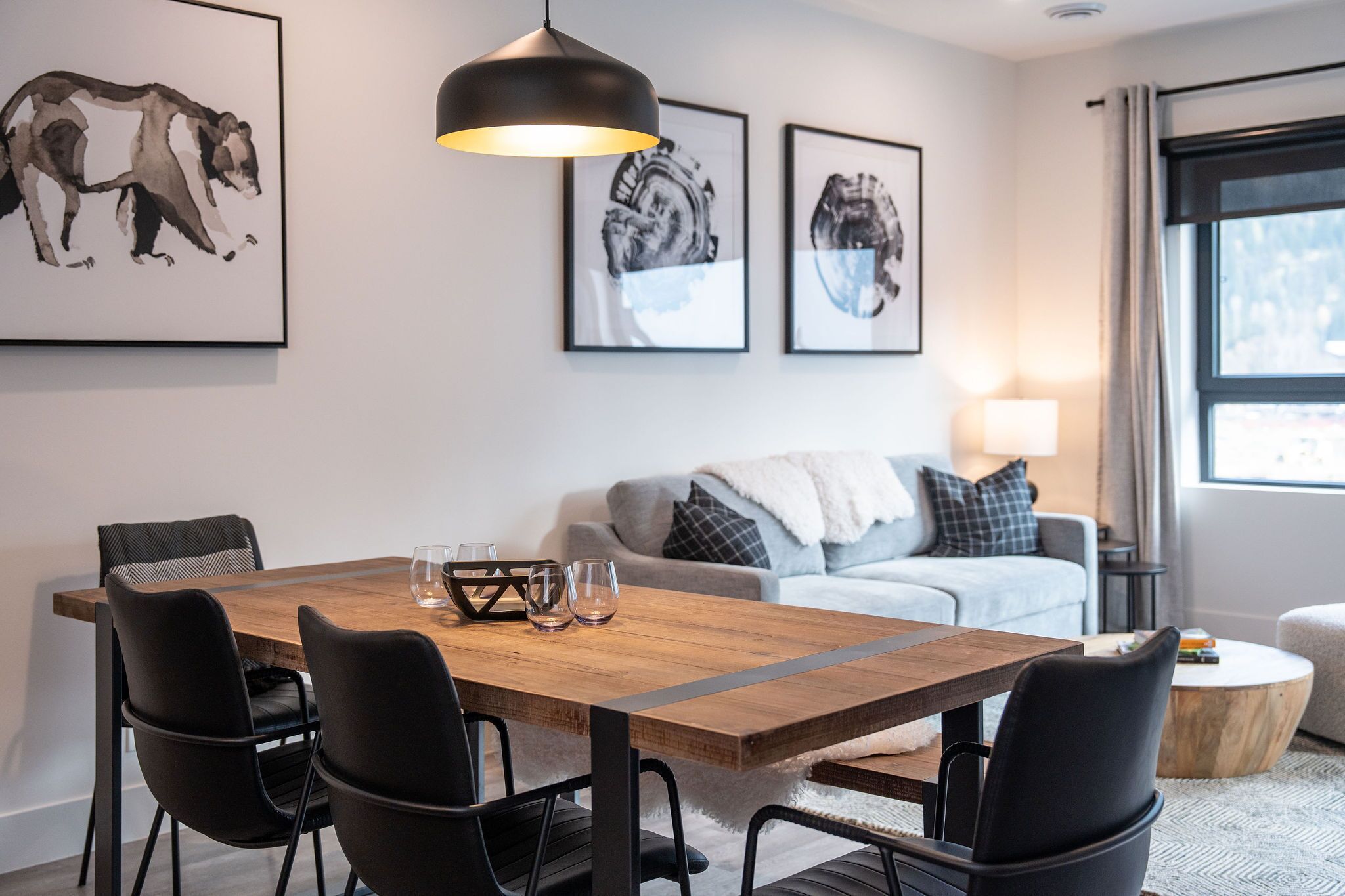A modern living room with a wooden dining table, black chairs, a grey sofa, and a large window with curtains.