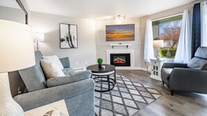 A modern living room with a grey sofa, a round coffee table, a fireplace, and a large window with curtains.