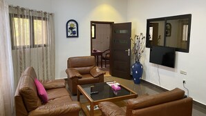 A living room with a brown leather sofa, a glass coffee table, a TV mounted on the wall, and a vase with dried flowers. - Cozyapartment (Cotonou)