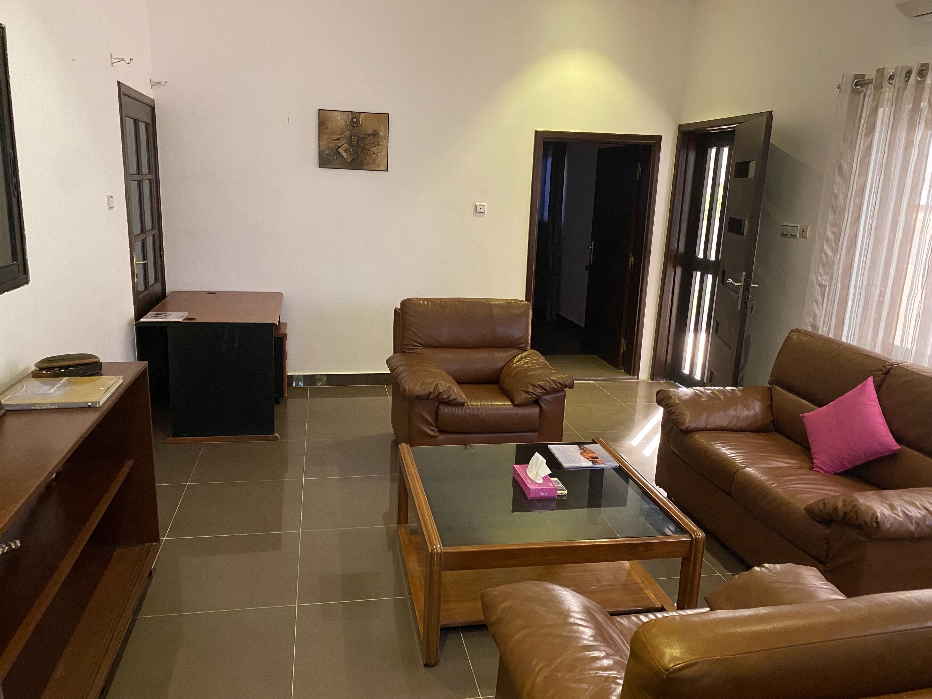A living room with a brown leather sofa, a glass coffee table, and a wooden side table. There is a door leading to another room and a wall-mounted artwork.