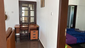 Quarto compacto com escrivaninha de madeira, uma cadeira, cama com cobertor azul e mesa de cabeceira pequena.