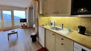 Une cuisine compacte avec des meubles en bois, un évier et un four à micro-ondes. On y trouve également une petite table et un téléviseur dans le salon attenant.