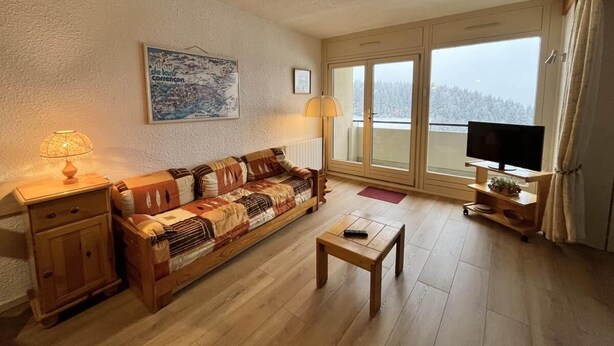 Un salon chaleureux avec un canapé en bois, une table basse, un téléviseur posé sur un meuble en bois et une grande fenêtre donnant sur les arbres.
