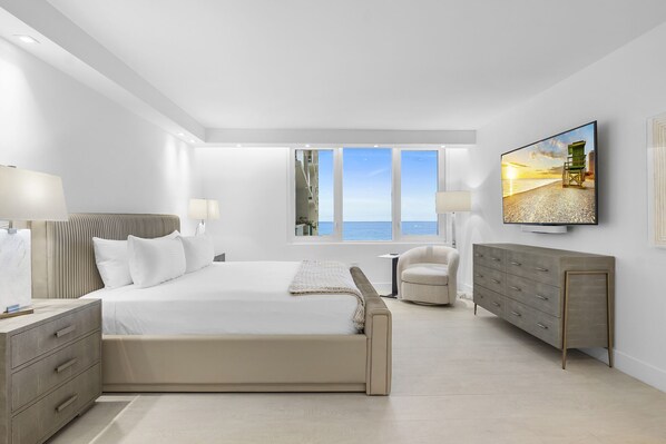 A modern hotel room with a large bed, a TV, a dresser, and a view of the sea.
