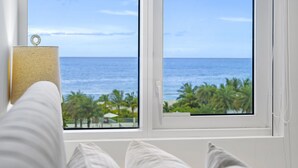 Ein Bett mit weißer Bettwäsche, ein Fenster mit Blick auf das Meer und Palmen sowie eine weiße Wand mit einer Tür.