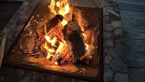 A square fire pit with burning logs, surrounded by stone tiles, on a wooden deck with two Adirondack chairs. - The Henhouse at The Yellow Barn Estate (Freeville)
