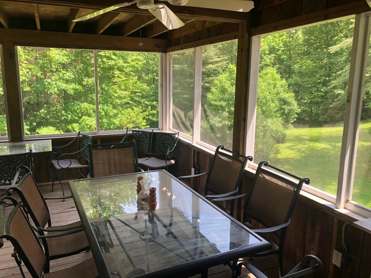 Een overdekt terras met een glazen tafel en stoelen, grote ramen en uitzicht op een groen landschap.