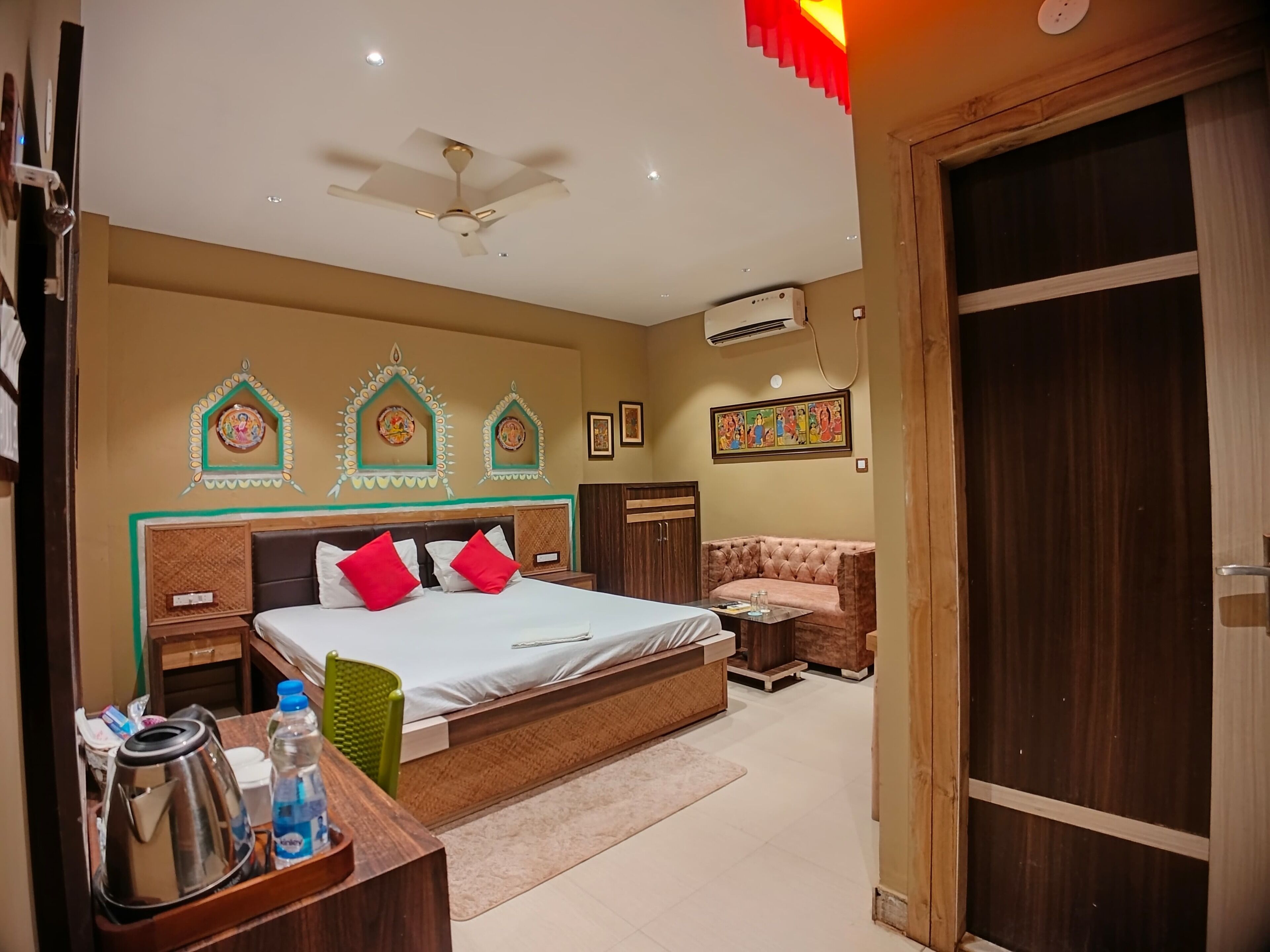 A hotel room with a bed, bedside tables, a desk with a kettle and water bottles, a ceiling fan, and decorative wall art.