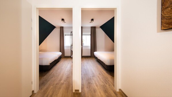 A mirrored room with two beds, wooden flooring, and a window with curtains.