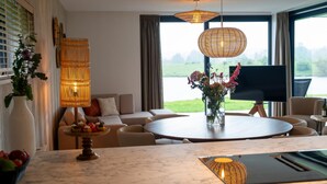 A modern living room with a round wooden table, a sofa, and a TV. There are two hanging lamps and a vase with flowers on the table. - Premium Island 6p in Thorn (Thorn)