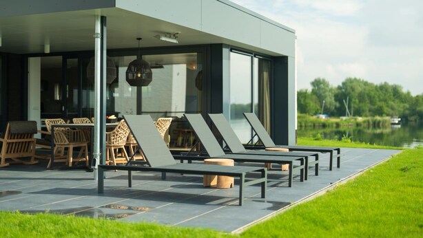 A modern outdoor seating area with lounge chairs and wooden chairs, a covered patio, and a view of a lake. - Premium Island 6p in Thorn (Thorn)