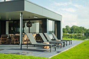 A modern outdoor seating area with lounge chairs and wooden chairs, a covered patio, and a view of a lake. - Premium Island 6p in Thorn (Thorn)