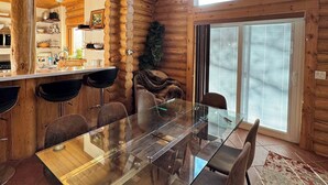 L’intérieur d’un chalet en bois comprend une table à manger en verre, des chaises, un coin cuisine et une grande fenêtre.