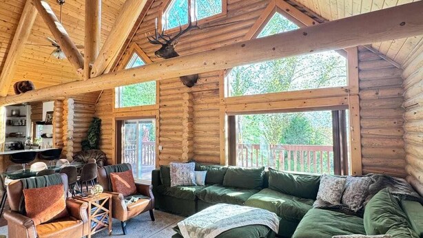 L’intérieur d’une cabane en rondins, avec un plafond haut, des poutres en bois, une cheminée, des meubles recouverts d’un tissu vert et un coin repas.