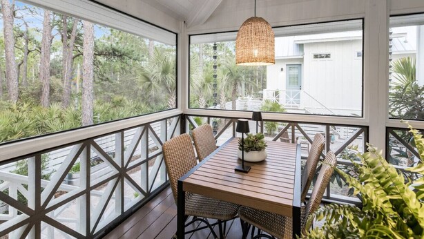 A covered patio with a wooden table and wicker chairs, overlooking a garden and a building. - Mahi Mahi Cottage in the Woods (Santa Rosa Beach)