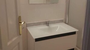 Une salle de bain avec un meuble-lavabo blanc, un miroir avec éclairage intégré et une porte dotée d’une poignée dorée.