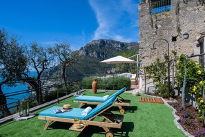 Enjoy breathtaking views of the coastline and mountains from this rooftop terrace, featuring lounge chairs, a shaded area, and a serene garden.