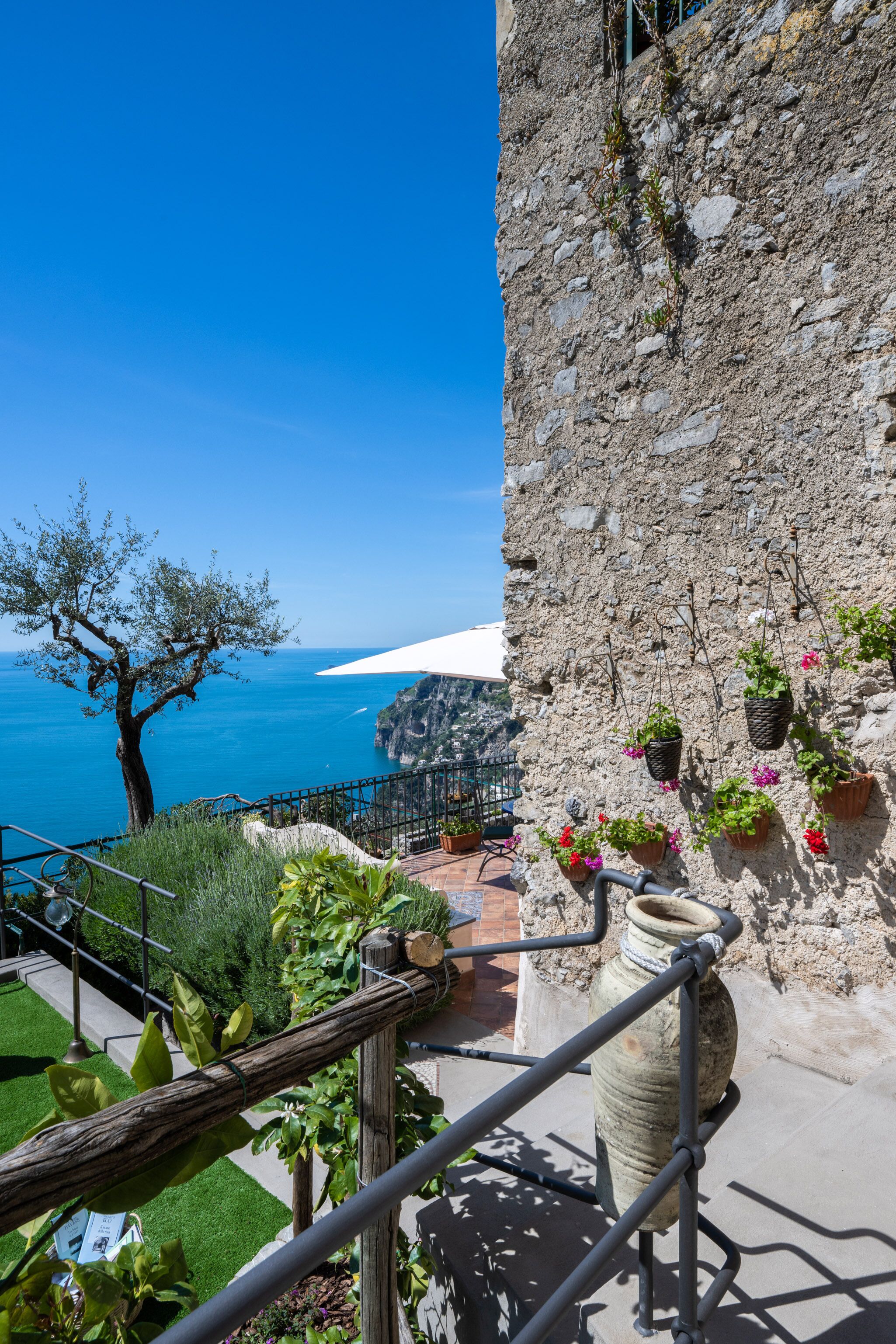 Un bâtiment en pierre offrant une vue sur la mer, agrémenté de plantes en pot et doté d’une rambarde en métal.