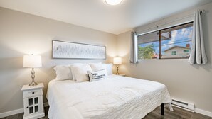 Une chambre à coucher avec un grand lit, deux tables de chevet avec des lampes, une fenêtre avec des rideaux et une œuvre d’art accrochée au mur.
