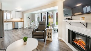 Une cuisine moderne avec des meubles blancs, un coin repas avec une table et des chaises blanches, un fauteuil en cuir et une cheminée avec des bûches.