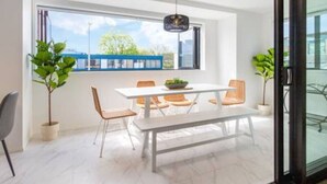 A modern dining area with a white table, orange chairs, and a bench. There are potted plants and a view of the outdoors through large windows.