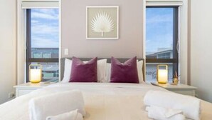 A hotel room with a large bed, two purple pillows, a framed artwork on the wall, and a window with a view of buildings.