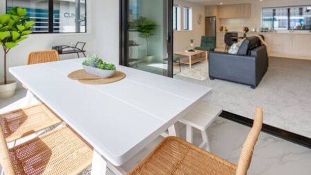 A modern living room with a white table, wicker chairs, a grey sofa, and a glass door leading to a kitchen.