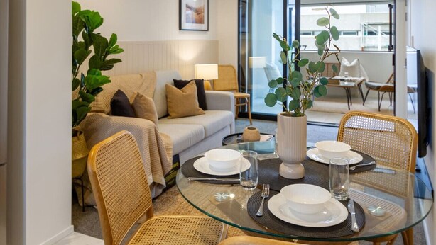 A dining area with a glass table set for four, surrounded by wicker chairs. A large plant is in the center, and a sofa with cushions is in the background. - Modern CBD Central Stay - Hagley Park + Balcony (Christchurch)