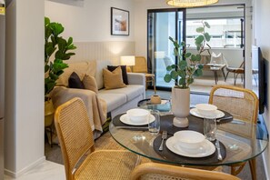Un comedor con una mesa de vidrio dispuesta para cuatro personas, rodeada de sillas de mimbre. Una planta grande está en el centro, y un sofá con cojines se observa al fondo.