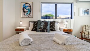 A neatly made bed with a pillow featuring a dog, a window with a view, and a chair with a patterned cushion. - Okioki - Seaside Escape (Christchurch)