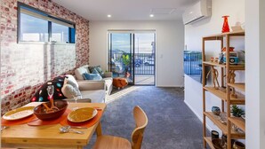 A modern living room with a brick wall, a sofa, a dining table set with plates and bowls, and a large window overlooking a balcony. - Okioki - Seaside Escape (Christchurch)