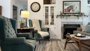 A living room with a green armchair, a side table with a lamp, a fireplace with a mantel decorated with books and greenery, and a clock on the wall. - Creekside Cottage Near Nashville- Pet Friendly! (Pegram)