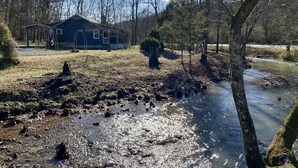 A rustic cabin with a porch, surrounded by trees and a small stream. - Creekside Cottage Near Nashville- Pet Friendly! (Pegram)