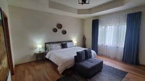 Une chambre à coucher comprenant un lit, des tables de chevet, une commode, un canapé, une fenêtre avec des rideaux et des éléments de décoration murale.