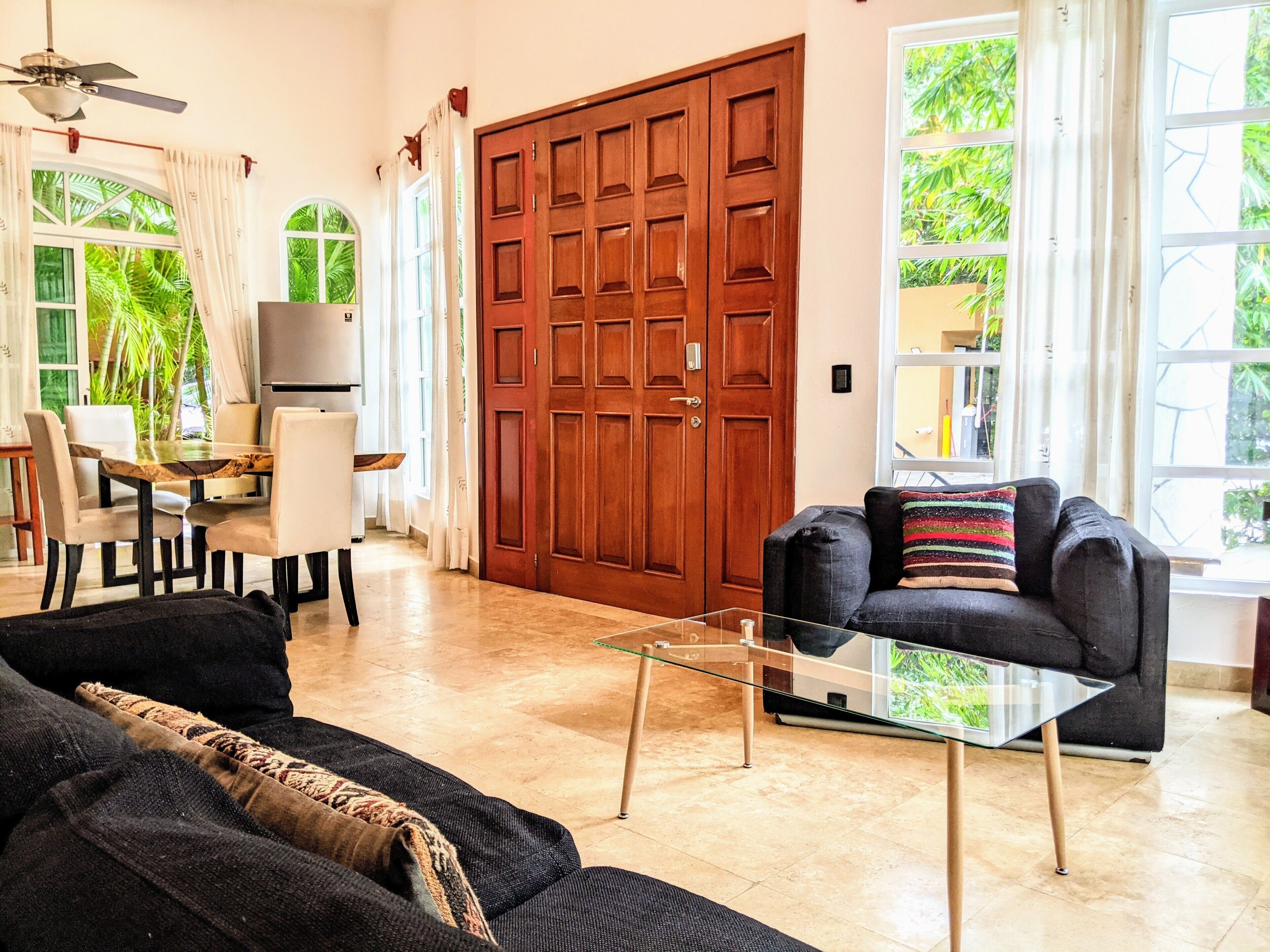 Ein geräumiges Wohnzimmer mit einer großen Holztür, einem Glastisch und einem schwarzen Sofa mit farbigem Kissen.