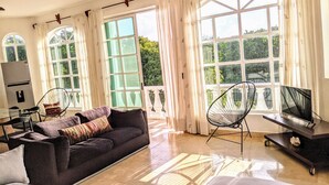 A spacious living room with a dark sofa, a TV on a stand, and large windows with curtains. - 9 Bed 6bath  Pool, Roof lounge, Beach Best location in Playa Walk to everything (Playa del Carmen)