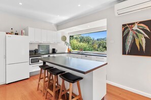 A modern kitchen with a large island, white cabinetry, and a view of greenery outside. - Coastal Charm (Lorne)