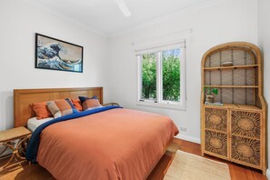 Un dormitorio con cama, ventana, mueble de ratán y una obra de arte enmarcada en la pared.