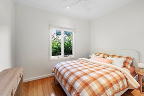 A bedroom with a bed, a window, a ceiling fan, and a wooden nightstand. - Coastal Charm (Lorne)