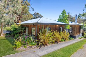 A modern house with a metal roof, wooden siding, and a well-maintained garden with various plants and flowers. - Coastal Charm (Lorne)