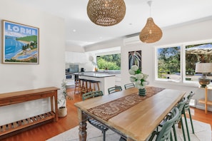 A modern kitchen with white cabinets, a dining area with a wooden table and chairs, and a large window with a view of greenery. - Coastal Charm (Lorne)