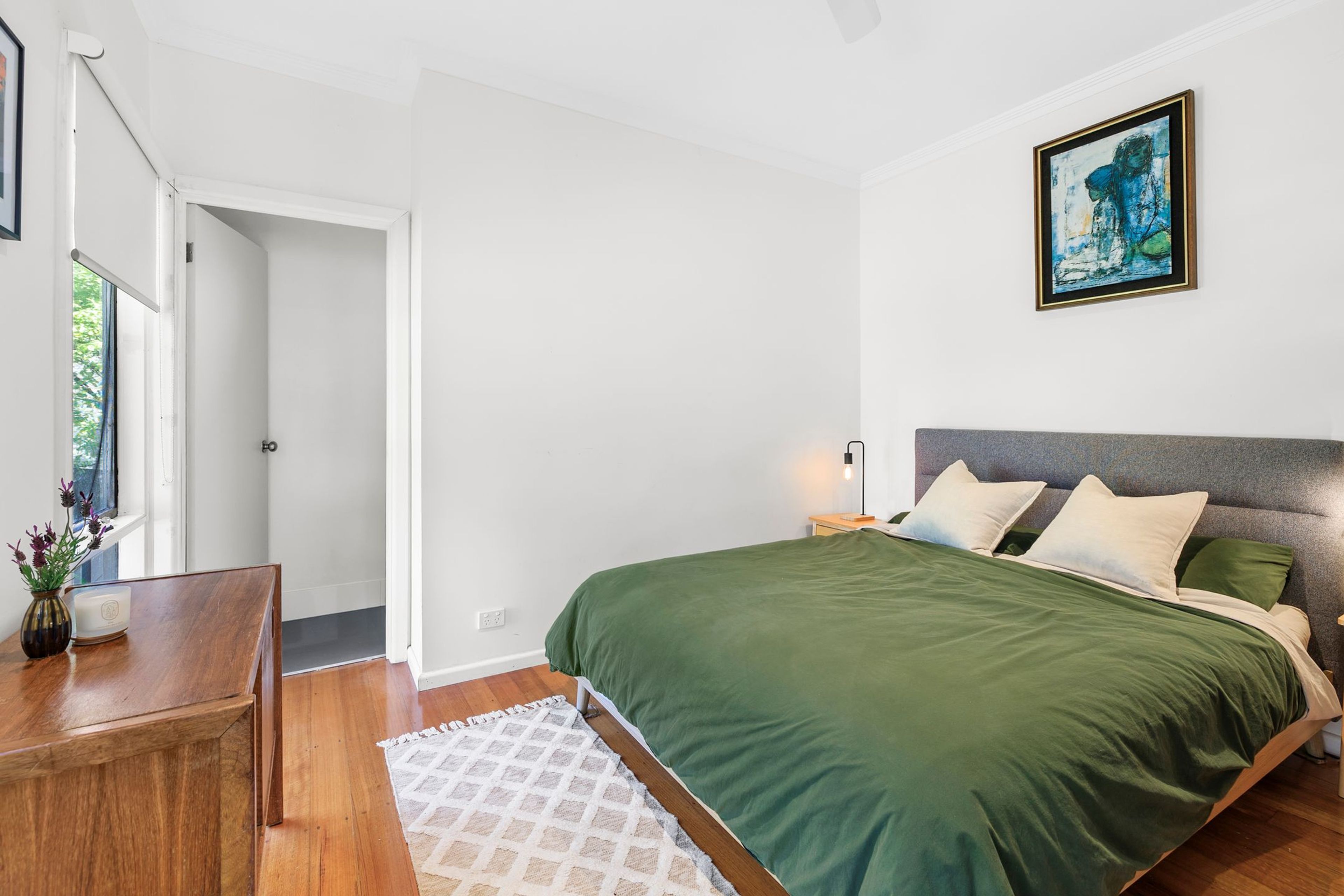 A bedroom with a bed, a nightstand, a bedside lamp, a painting on the wall, and a door leading to another room.