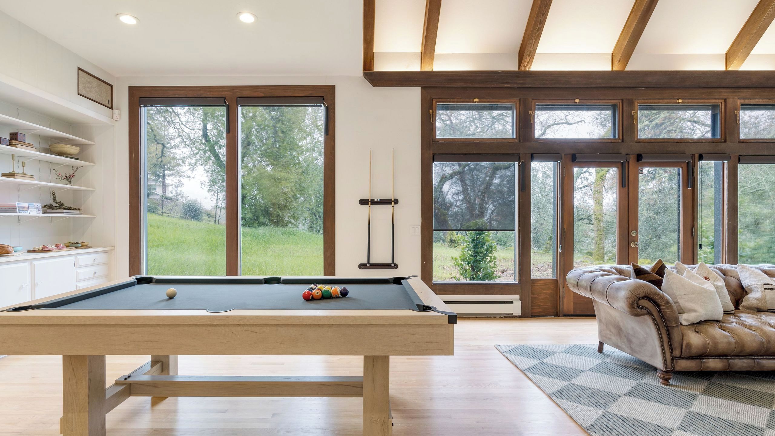 Une table de billard dans une pièce dotée de grandes fenêtres, d’un canapé et d’une bibliothèque.