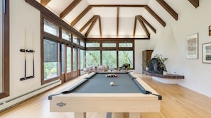 A pool table in a room with large windows, wooden beams, and a fireplace.
