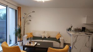 A modern living room with a sofa, coffee table, and two chairs. There's a large window, a plant, and a kitchen area with a marble countertop. - Maison Neuve Bord de mer (Lion-sur-Mer)