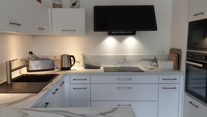 A modern kitchen with white cabinetry, a marble countertop, a built-in oven, and a ceiling fan. - Maison Neuve Bord de mer (Lion-sur-Mer)